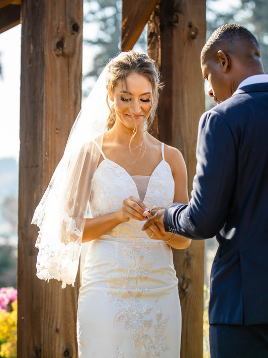 Bride in an elegant white lace wedding dress and groom in a sharp dark suit exchanging rings under a wooden arbor, capturing a heartfelt moment of their outdoor wedding ceremony – perfect attire inspiration for Portland, OR weddings