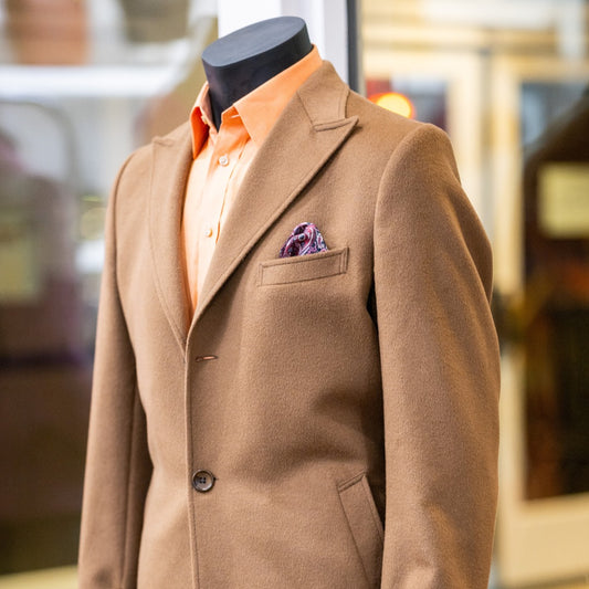 Close-up view of a custom-tailored camel brown coat with a vibrant orange shirt and a patterned pocket square, designed by the House of Rose, displayed on a mannequin